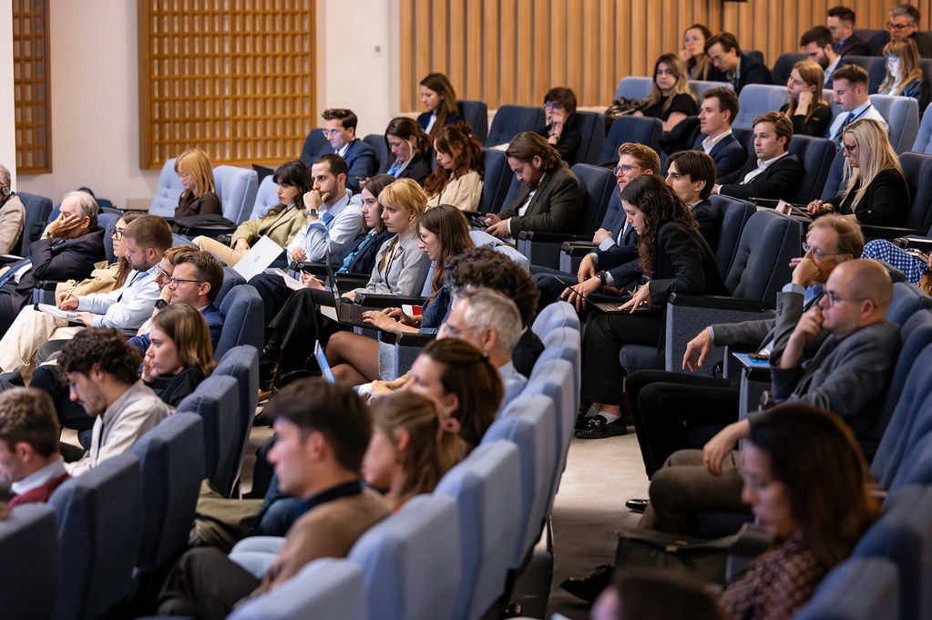 Audience seated in a modern lecture hall, many taking notes or looking at devices during a conference session on stage left/right of image.