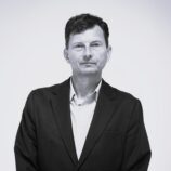 Head-and-shoulders portrait of a man in a dark suit jacket and white shirt against a light gray background, looking at the camera with a subtle smile.