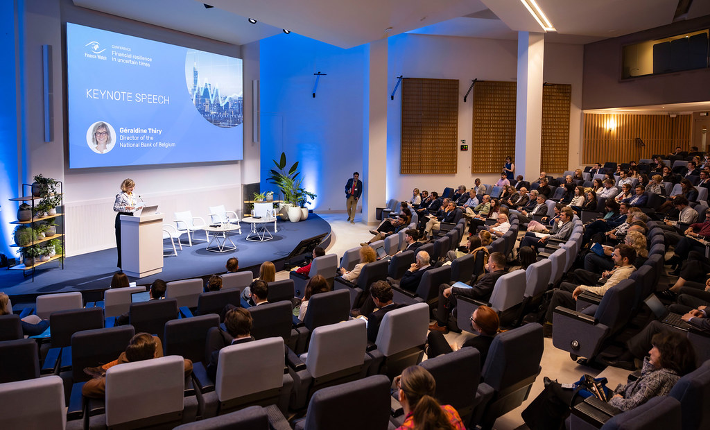 Speaker at a podium presenting a keynote on a blue-lit stage to a large seated audience in a conference hall.