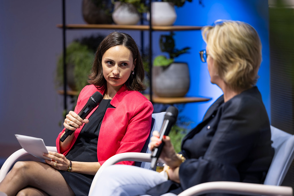 Two women in a studio interview, microphones in hand; left woman in a pink blazer with papers, right woman in a dark blazer speaking, blue backdrop with shelves behind them.