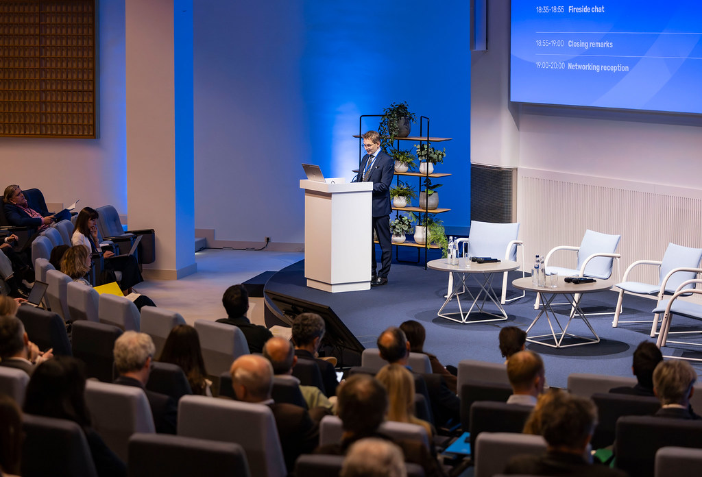 Male presenter in a suit speaks at a white podium on a blue-lit conference stage, with potted plants on a shelving unit behind him and an audience in seating to the left and foreground.