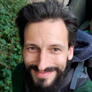 Close-up of a man with dark hair and a full beard smiling, outdoors with green plants in the background and a backpack strap visible.