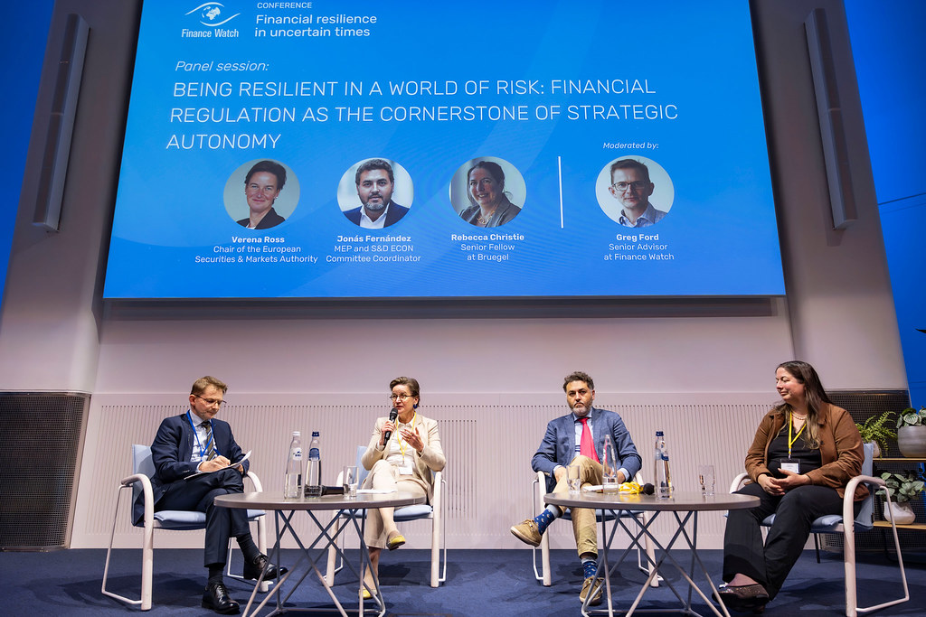 Panel discussion at a conference with four speakers seated on stage beneath a large blue screen showing the title: 