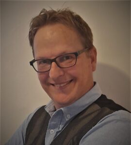 Smiling man with short light brown hair and glasses wearing a blue button-down shirt and dark patterned vest, posing for a portrait in a neutral setting.