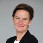 Professional head-and-shoulders portrait of a smiling woman with short brown hair, wearing a black blazer and white blouse against a gray background, arms crossed.