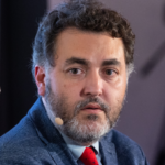 Close-up of a bearded man in a blue blazer and red tie speaking at a panel, wearing a microphone headset.