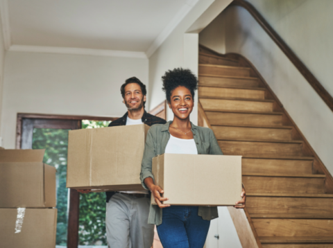 Two people carrying cardboard boxes inside a home near a wooden staircase, smiling as they move in together.